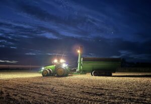 A green tractor and a green grain cart sit parked on a field of stubble from a harvested soybean field. The sky is a moody dark blue and the lights on the tractor and grain cart are shining bright against the dark sky. 