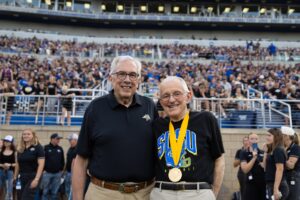 Two men stand shoulder to shoulder in front of the stadium seats of a football stadium. The stadium is full of spectators and the two men are both wearing black shirts. The man on the right wears a gold medal hanging from a yellow ribbon around his neck. 