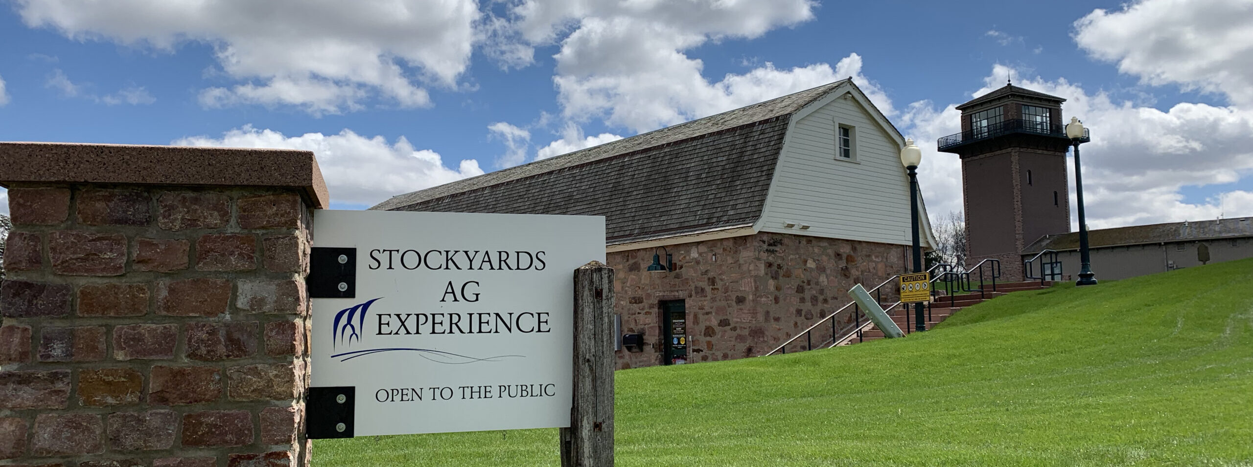 The stone sign for the Stockyards Ag Experience, the historic quartzite pink and white wood museum barn, the visitor's towner on day with blue skies and green grass