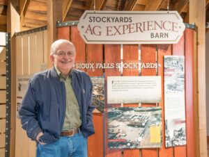 An older gentleman stands by a wooden museum display. He's smiling and wearing a smile outfit of jeans, a light olive green shirt and a navy blue jacket. 