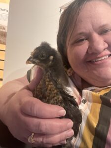 a woman with short hair and a bright striped shirt holds a baby chick that is brown and black